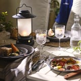 Set da tavola con porcellana floreale moderna, STRIMMIG. La lanterna centrata fornisce una luce calda. I piatti contengono cibo fresco. Bicchieri e decanter suggeriscono bevande.
