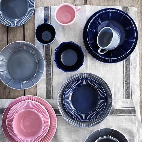 Stoviglie STRIMMIG blu, rosa, grigie con disegni floreali su tovaglia a righe.