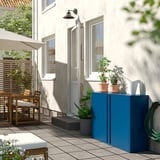 Piccolo balcone con armadietto verde acqua, piante in vaso, tavolo in legno e posti a sedere all'aperto bianchi sotto un edificio beige.