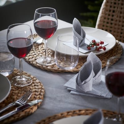 Tavola apparecchiata con grandi bicchieri da vino, piatti, posate e tovagliette intrecciate.