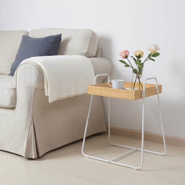 Tavolino in bambù bianco SVENARUM accanto al divano beige, con fiori e una tazza blu, che aggiunge un tocco chic minimalista.