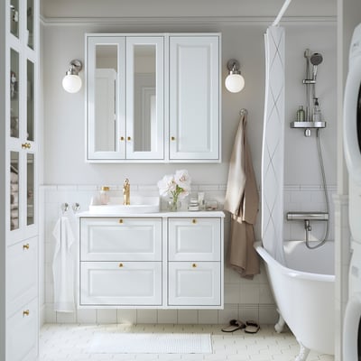 Bagno bianco moderno con mobile TÄNNFORSEN, accenti dorati, mensola in vetro e fiori. Mobile poco profondo sopra il lavandino, che si adatta bene tra le applique.