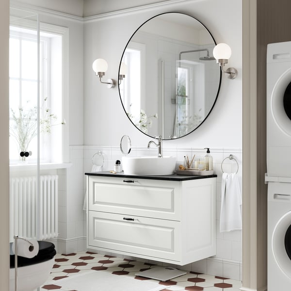 Bagno moderno bianco con mobile TÄNNFORSEN. Specchio rotondo, lavello a vaso, piastrelle geometriche per pavimenti, lavatrice bianca. Design elegante e spazioso.