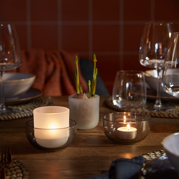 Set da tavolo in legno con lumini accesi in portabicchieri, bicchieri da vino, piatti e piantine in vaso.