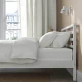 Un letto di legno bianco vicino a una finestra, ben fatto con lenzuola e cuscini bianchi. Sopra è montata una lampada da parete.