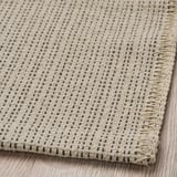 Tappeto in cotone a tessitura piatta, beige con piccoli punti scuri, ben bordato, posato su pavimento in legno.