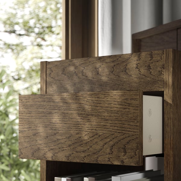 Cassettiera in legno scuro dal design moderno e cassetti con chiusura ammortizzata, posizionata in prossimità di una finestra con vista verde sull'esterno.