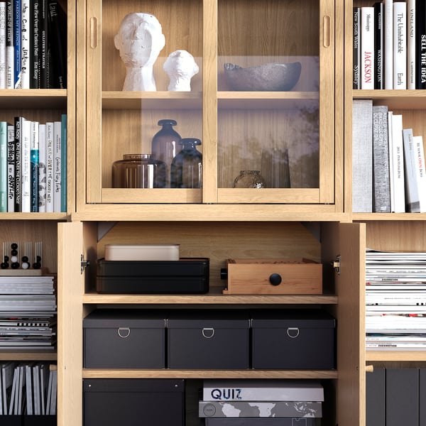 Libreria in legno con ante in vetro, che espone oggetti decorativi e libri. Contenitori a giorno e chiusi, ripiani regolabili e maniglie integrate.