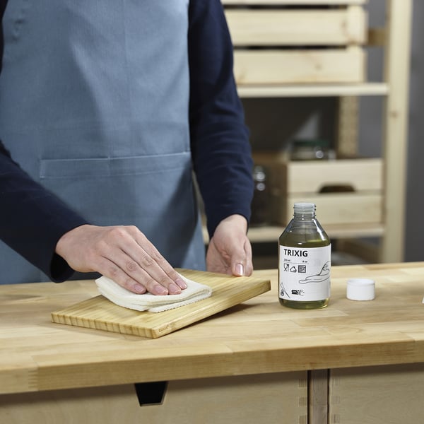 La persona applica l'olio sulla tavola di legno con un panno. Bottiglia etichettata TRIXIG nelle vicinanze.
