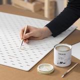 Una persona sta applicando la vernice su un pannello bianco usando un pennello. Una lattina etichettata TRIXIG e un piccolo contenitore di vernice si trovano nelle vicinanze sul tavolo.