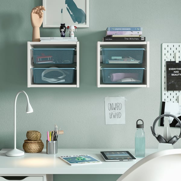 Scaffali TROFAST bianchi con scatole blu contenenti libri e decorazioni. Scrivania con lampada, tablet e bottiglia d'acqua sottostante.