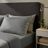 Accogliente camera da letto con lenzuola grigie ULLVIDE, testiera grigio scuro e tocchi personali sul comodino.