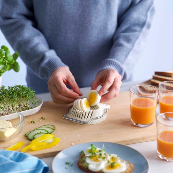 Persona che prepara fette di uovo sodo usando una pratica affettatrice per uova su una tavola di legno con vari prodotti per la colazione intorno.