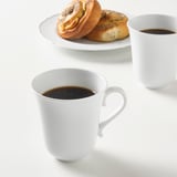 Due tazze bianche con caffè, una con manico, e pasta frolla su piatto. Le tazze sono in porcellana durevole e resistente agli urti.