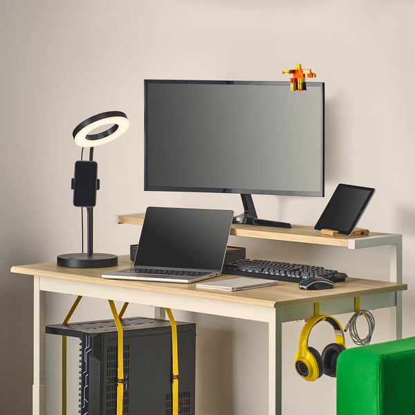 Una scrivania da gioco compatta con altezza regolabile, un ripiano per monitor e gestione dei cavi. Le cuffie sono appese lateralmente.
