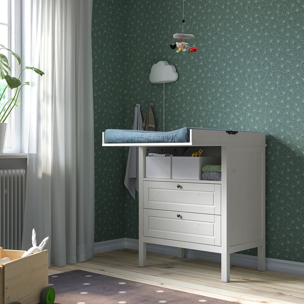 Nursery con carta da parati verde, fasciatoio bianco, coperte in cotone biologico, giocattoli e cestino in legno.