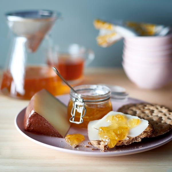 Un piatto con pane, formaggio, marmellata di arance e una brocca. VAGGSÅNG crema spalmabile all'arancia e ai fiori di sambuco.