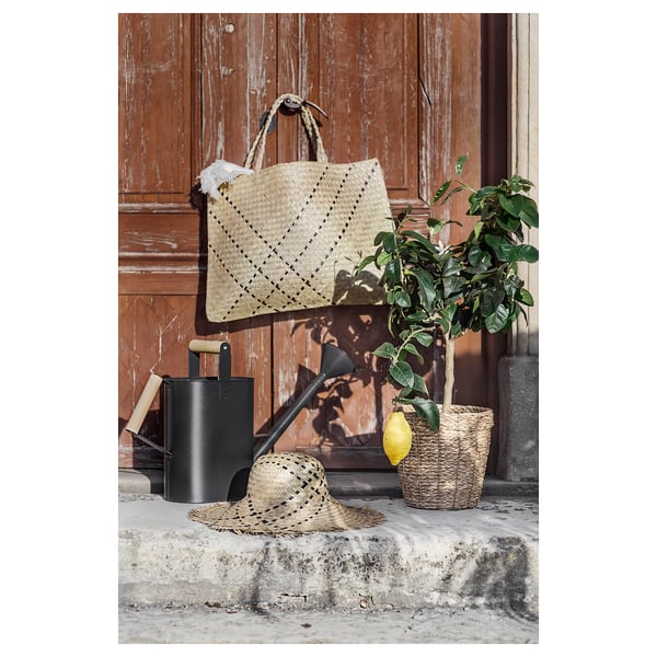 Cappello e borsa in erba marina intrecciata con pianta di limone e annaffiatoio contro porta in legno.