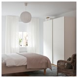 Camera da letto luminosa e minimalista con pareti bianche, copriletto grigio, lampada a sospensione bianca sospesa, sedia in vimini e grande finestra con tende.