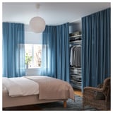 Elegante camera da letto moderna con tende blu VIDGA, biancheria da letto bianca, lampada bianca sospesa e sedia in vimini.