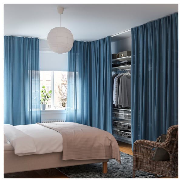 Camera da letto con tende VIDGA blu, biancheria da letto bianca, vestiti appesi, sedia di vimini, pianta e lanterna di carta.