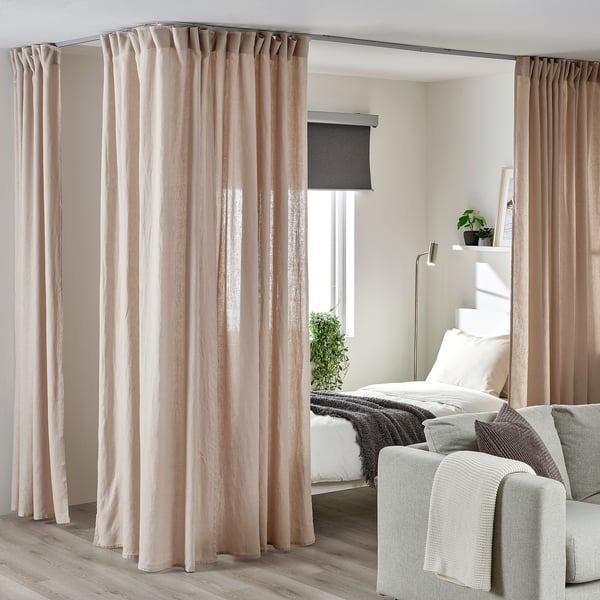 Camera da letto con tende VIDGA intorno a una grande finestra, configurazione accogliente. Letto bianco, divano, piante, pavimento in legno chiaro.