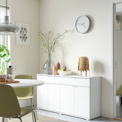 Moderna sala da pranzo minimalista con credenza VIHALS bianca, sedie verdi, fiori gialli in un vaso e un orologio rotondo.