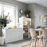 Spazio abitativo moderno con mobili contenitori bianchi VIHALS, sedie grigio chiaro, piante decorative e tappeto colorato.