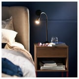 Comodino in noce VIKHAMMER con cassetto, mensole, lampada, libri e vetro accanto al letto.
