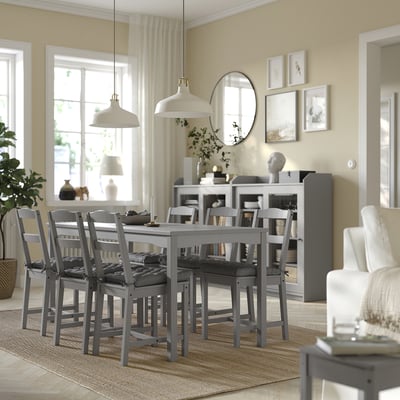 Sala da pranzo moderna con cuscini per sedie VIPPÄRT grigi, tavolo in legno, luci a sospensione, tappeto, tende, finestre, piante e decorazioni murali.