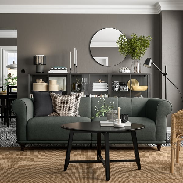 Soggiorno moderno con divano grigio scuro, cuscini fantasia, tavolino nero, lampada da terra e mensole decorate contro una parete grigia.