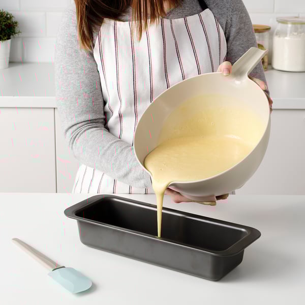 Persona che versa la pastella dalla ciotola inclinata con manico in una teglia rettangolare nera.