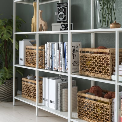 Scaffale in metallo bianco con vetro, che espone libri, cesti intrecciati e oggetti decorativi.