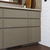 Cucina moderna con frontali dei cassetti lucidi grigio-marroni VOXTORP, piano di lavoro beige, accessori in legno e alzatina in stile mattone bianco.