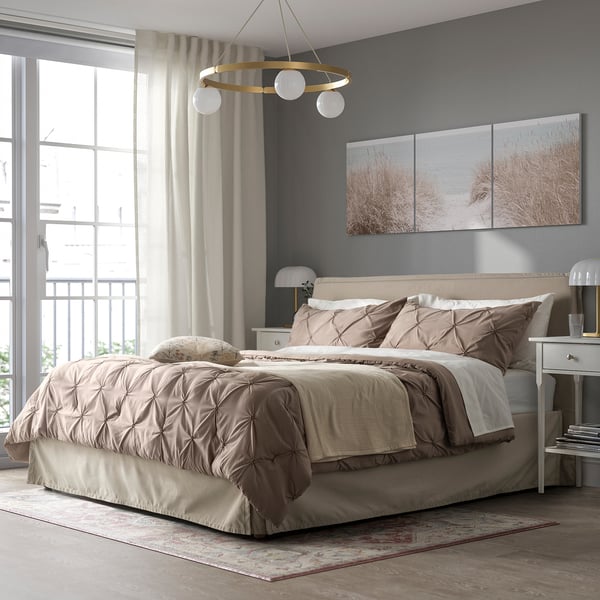 Cosy bedroom with beige BÄGARKLOCKA bedding, gold chandelier, grey walls & rug, beach prints, & bedside table lamps.