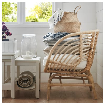 Natural woven BUSKBO armchair with cushions near a white table and window, offering a cosy, natural aesthetic.