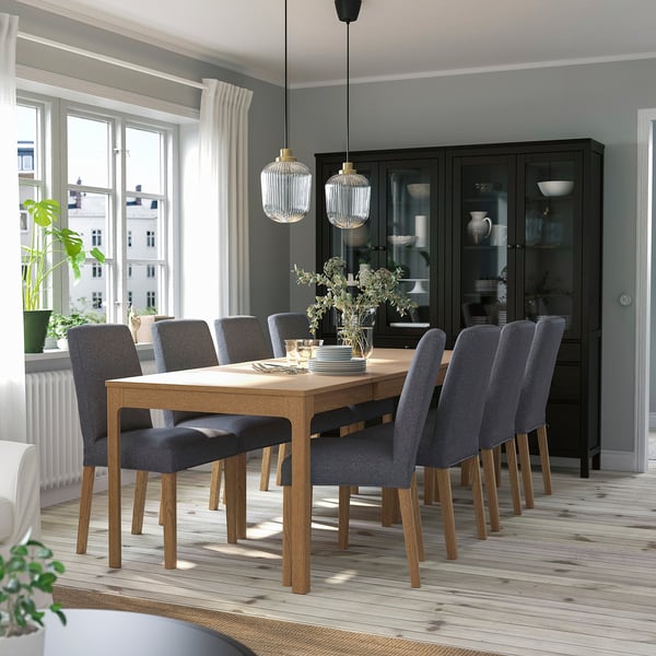 Modern dining room with grey upholstered chairs, wooden table, black cabinet, and large windows. Pendant lights hang above the table.