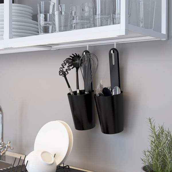 Black hanging utensil holders with tools below white shelves holding glasses and plates. Plant in front.