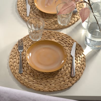 Two woven placemats with plates, cutlery, and glasses. Plates are simple, rustic, and somewhat smaller than the placemats.