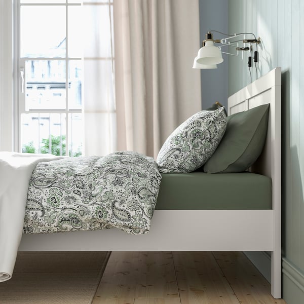 Bedroom scene with FÅGELFJÄLLET bed. Green pillows, white paisley quilt, wall lamp, and window view.