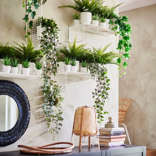 Indoor plants decor: white shelves with many FEJKA fake green plants, books, wicker lamp, and dark round mirror.