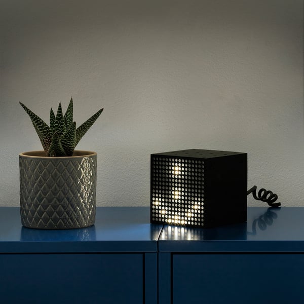 A black cube lamp with a textured planter holding a succulent sits on a blue shelf. The lamp responds to sound and has multiple light motifs.