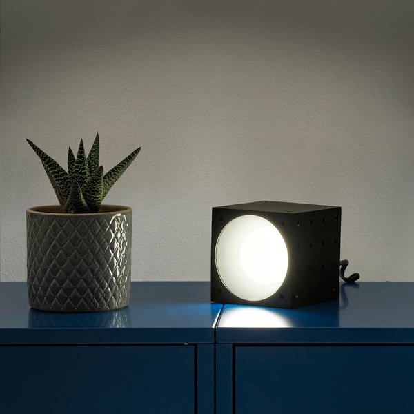 A modern table lamp beside a succulent plant on a blue shelf. The cube-shaped lamp emits a circular light.