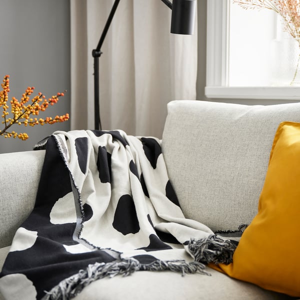 Living room with KLYNNETÅG throw on grey sofa, yellow pillow, modern lamp, and vase of orange berries. Natural light creates warmth.