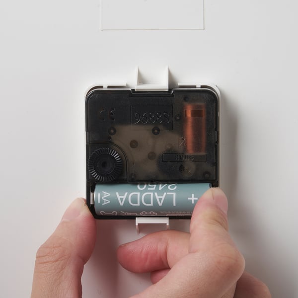 A person instals batteries into a square wall clock. The clock has a transparent case showing gears.