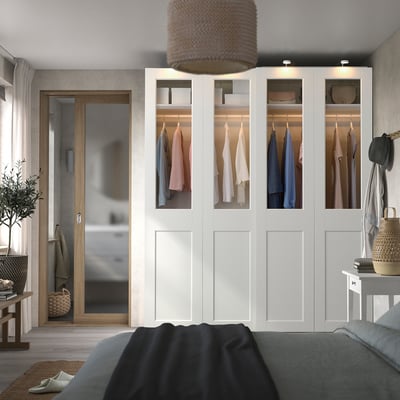 Modern bedroom with large white PAX wardrobe, see-through doors, hanging clothes, beside wooden door, black bed, wicker basket, and plant.
