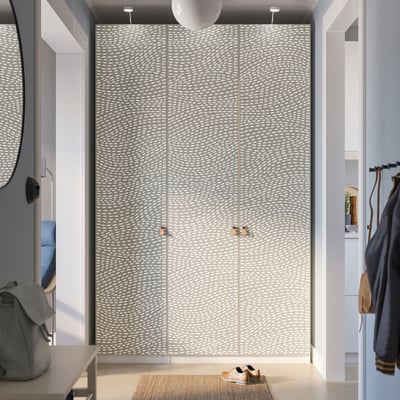 A modern wardrobe named PAX in a hallway. It white and grey wavy-patterned doors with gold handles. It stands tall next to a coat hanger and a mirror.