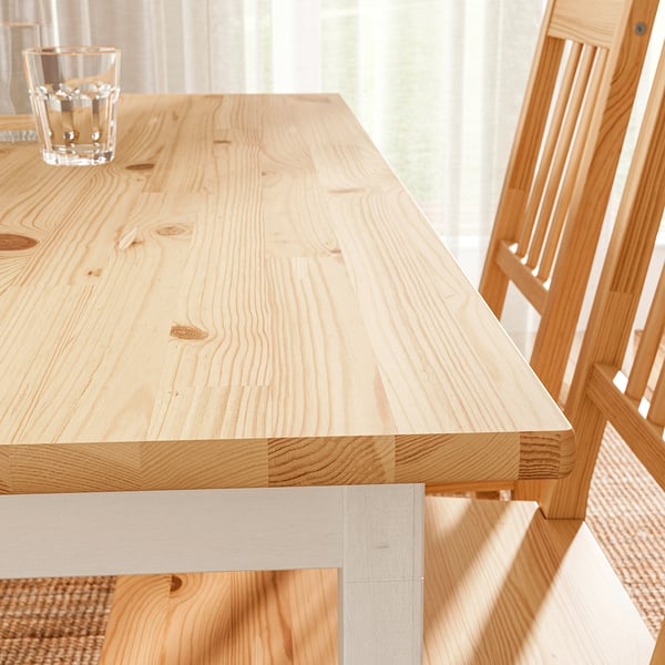 Wooden dining table with light finish, smooth top and sturdy legs on left side, next to chair with woven seat and backrest.