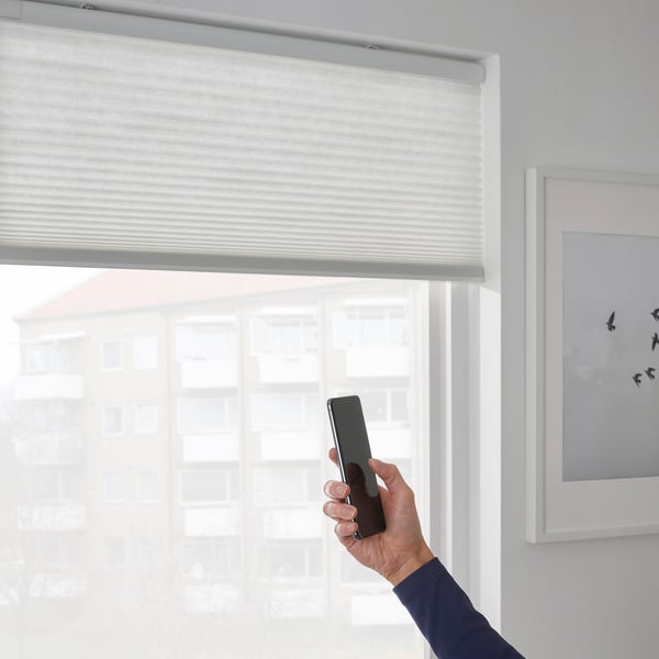 Hand holds smartphone controlling smart window blind using ikea home smart app, showing modern, safe, cordless design.