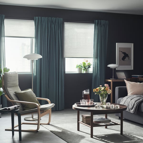 Modern living room with dark walls, teal curtains, wooden coffee table, grey armchair, and large window with blinds.